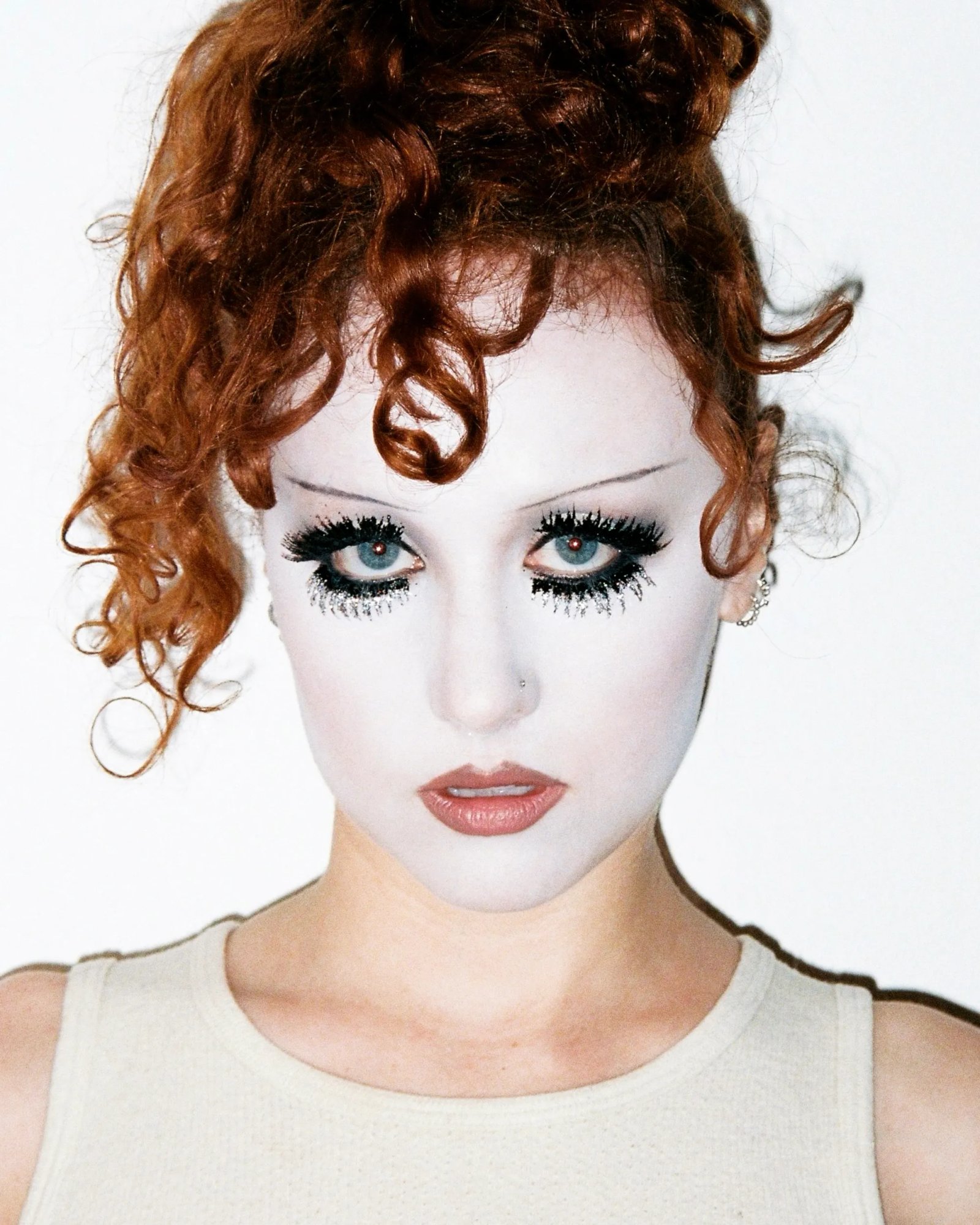Chappell Roan with white face makeup, drawn-on eyebrows, elaborate black and silver glitter eyelashes, a nose piercing, and curly red hair in a bun.