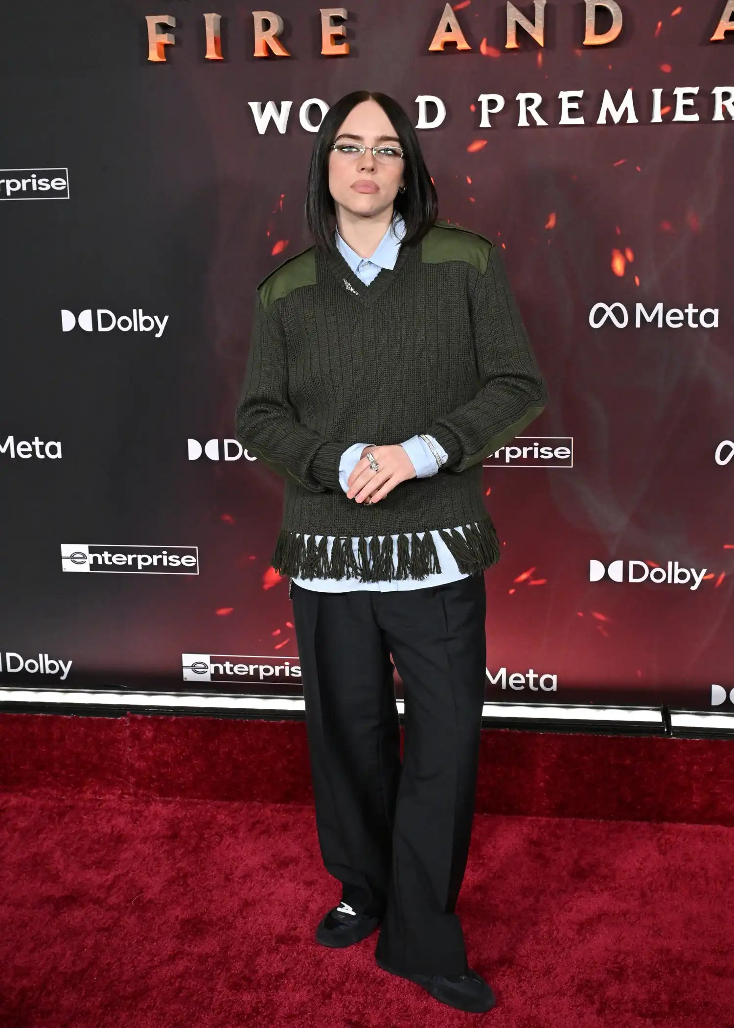 Billie Eilish wearing a green v-neck sweater with trousers and sneakers