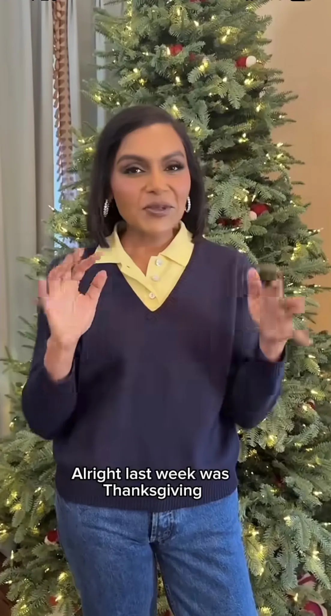 Mindy Kaling wearing a blue v-neck sweater with a yellow button-up and blue jeans
