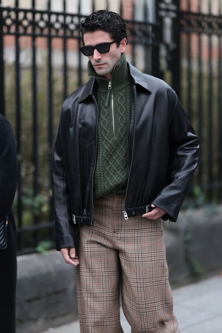 Model-turned-celebrity stylist Marc Forne at Paris Fashion Week, wearing a green cable-knit quarter-zip with a black leather jacket and brown plaid patterned trousers.