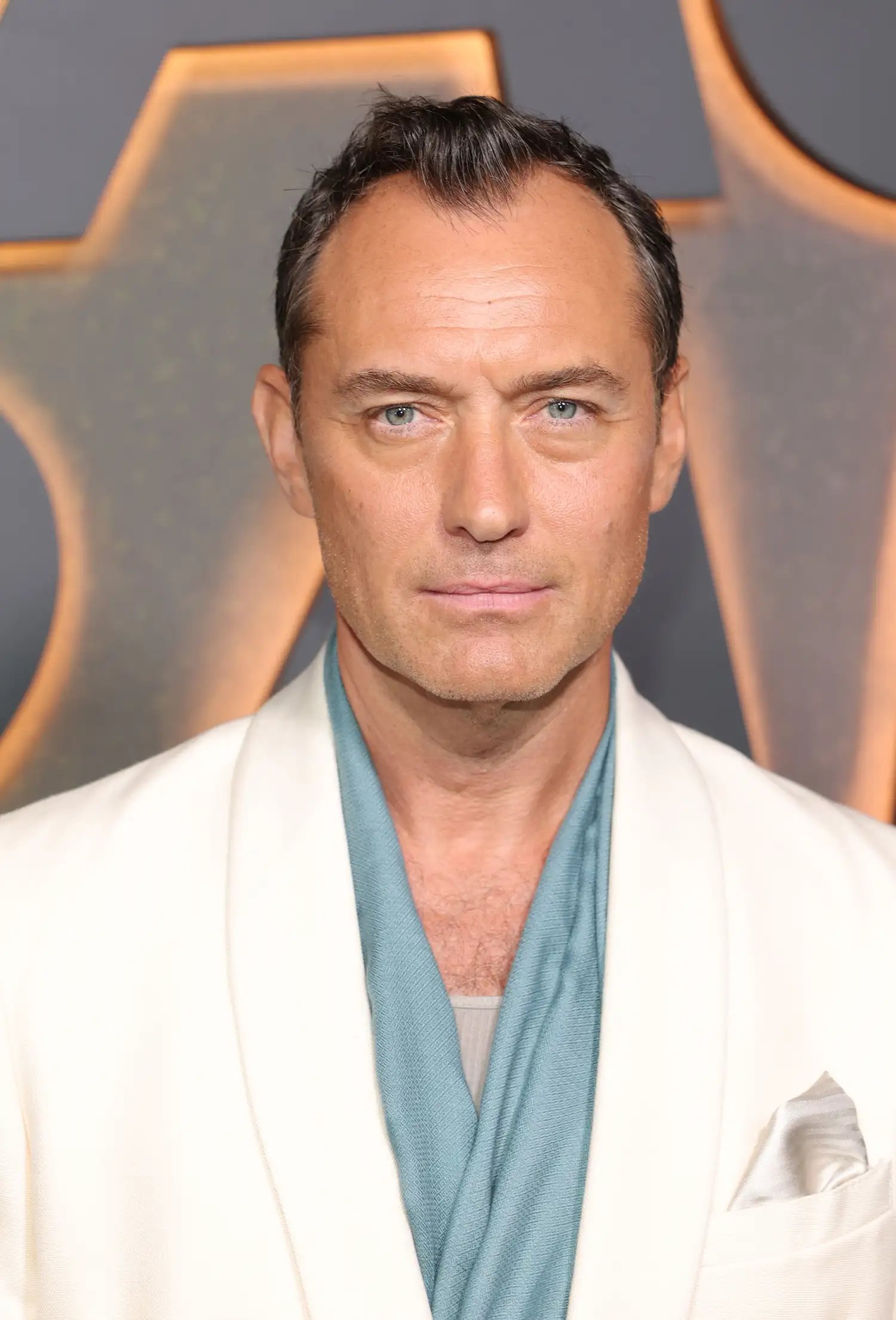 Jude Law with dark hair in a white blazer and blue scarf