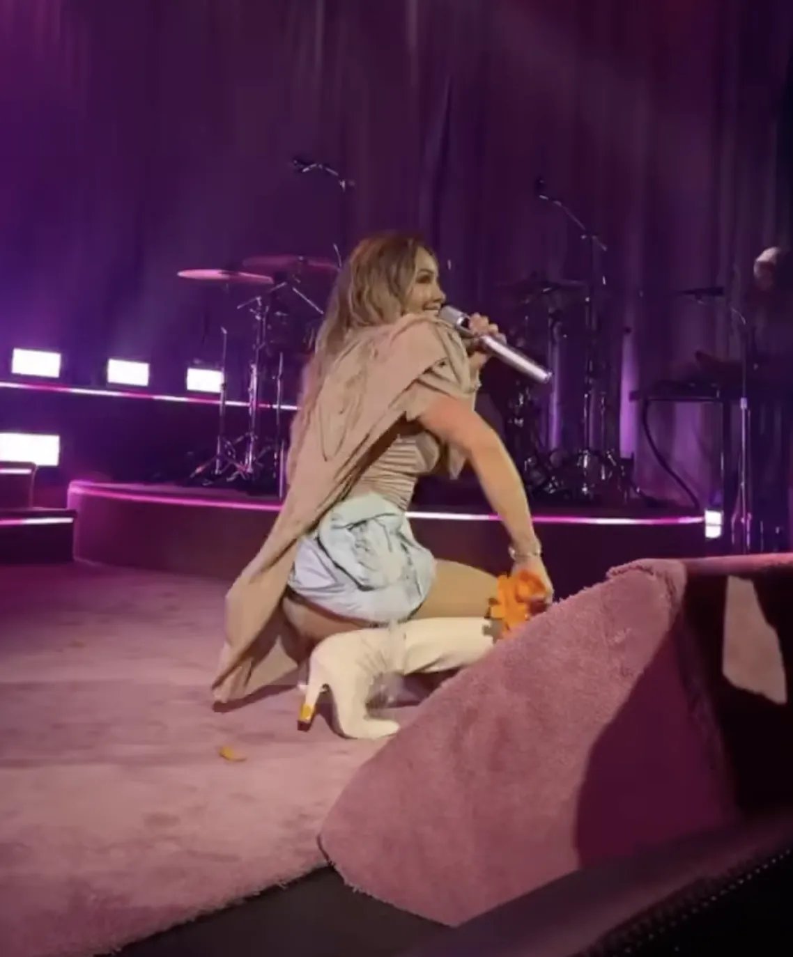 Hilary Duff in a nude bodysuit with a cape and rosette details singing onstage in London