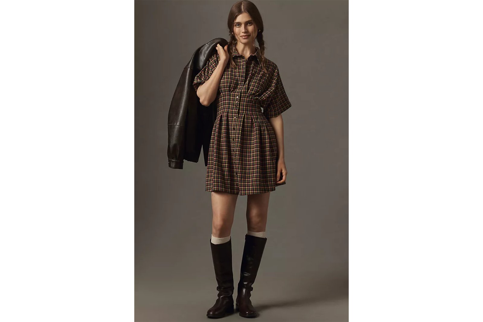 A model wearing a short-sleeved plaid dress with a dark leather jacket and knee-high boots.