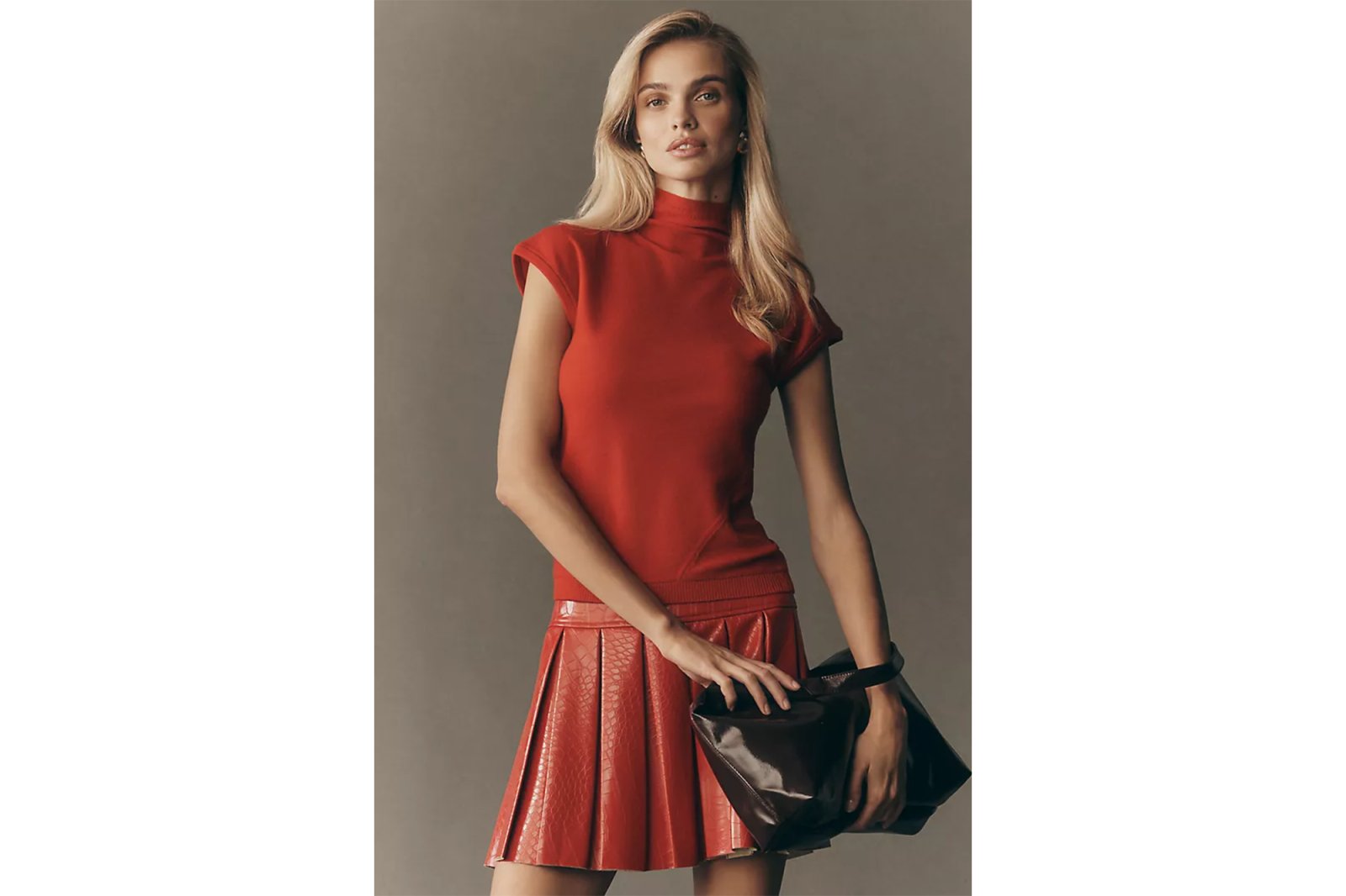 A woman in a red turtleneck top and pleated red skirt holding a dark brown bag.