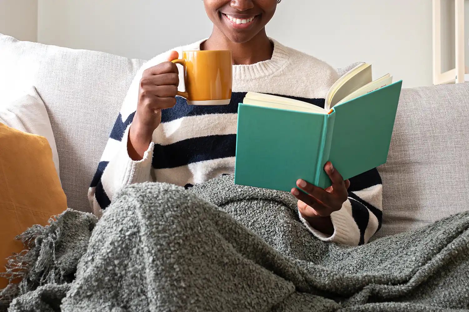 Stock image of an African American woman reading a novel at home, relaxing, drinking a cup of coffee.