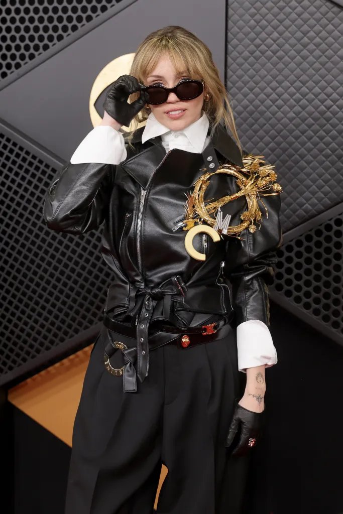 Miley Cyrus wearing a black leather jacket with a large gold brooch, black trousers, and sunglasses on the red carpet.