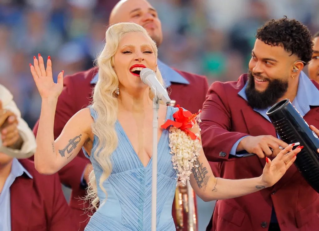 Lady Gaga singing into a microphone during a Super Bowl halftime show, wearing a light blue dress with red and white floral accents.