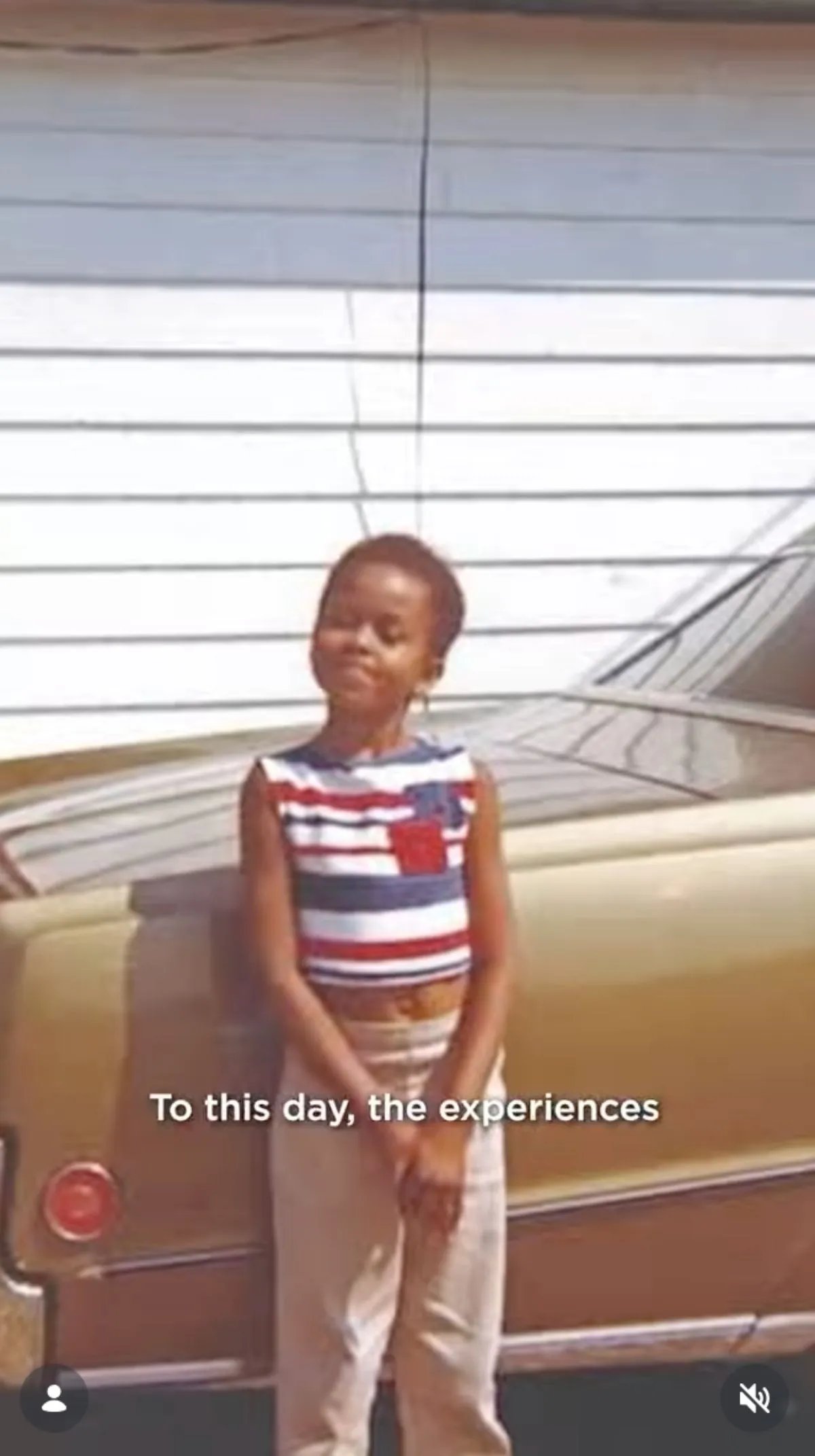 A grainy photo of Michelle Obama as a child. She is standing alone next to a car and smiling at the camera with her arms crossed in front of her. She's wearing a striped sleeveless shirt and trousers.