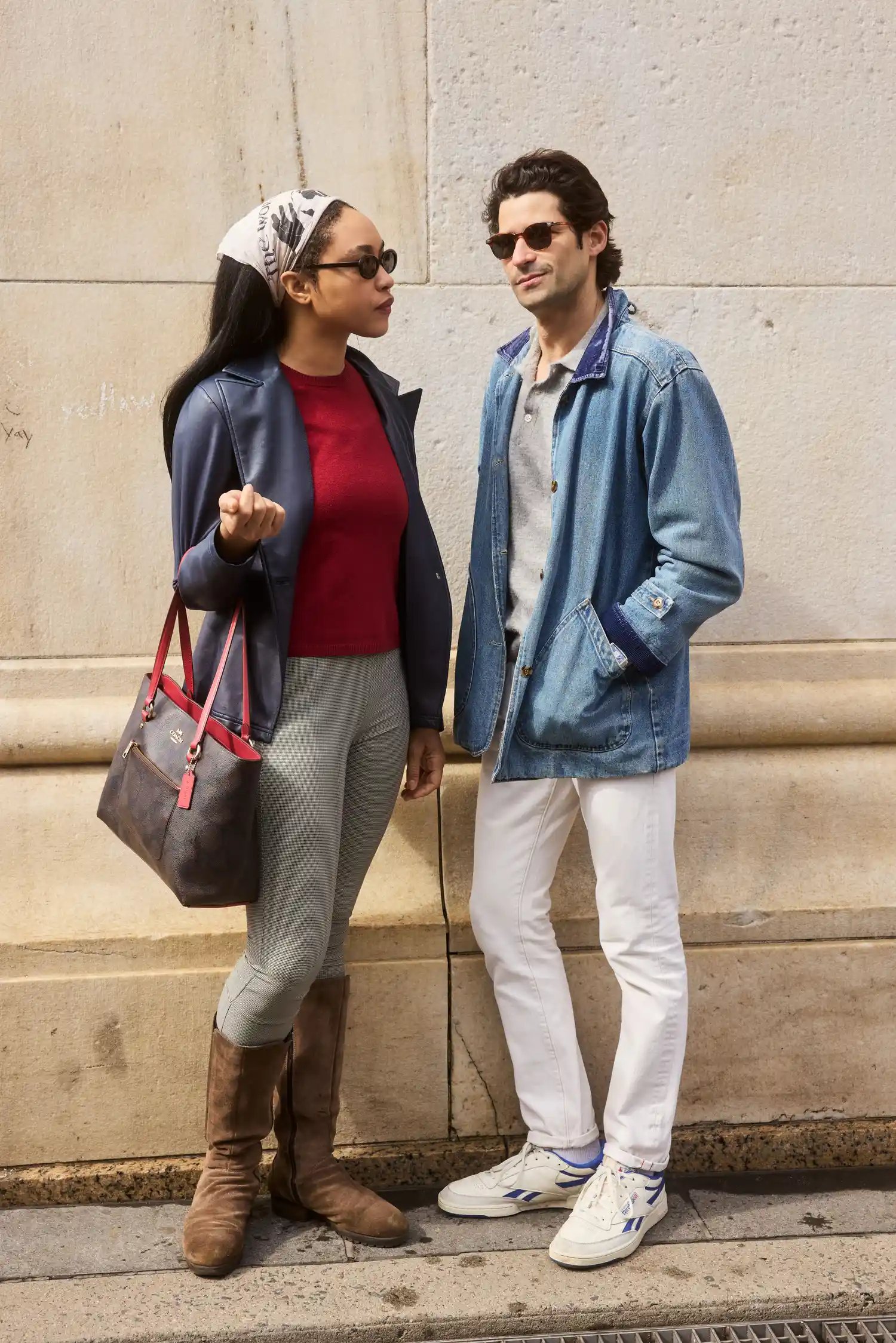 Two people standing on a sidewalk one dressed in casual chic attire carrying a handbag the other in light denim with sunglasses