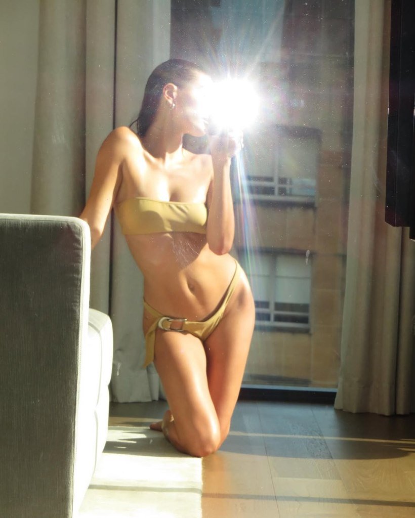 Hailey Bieber in a gold bikini, kneeling on a wooden floor in front of a window.
