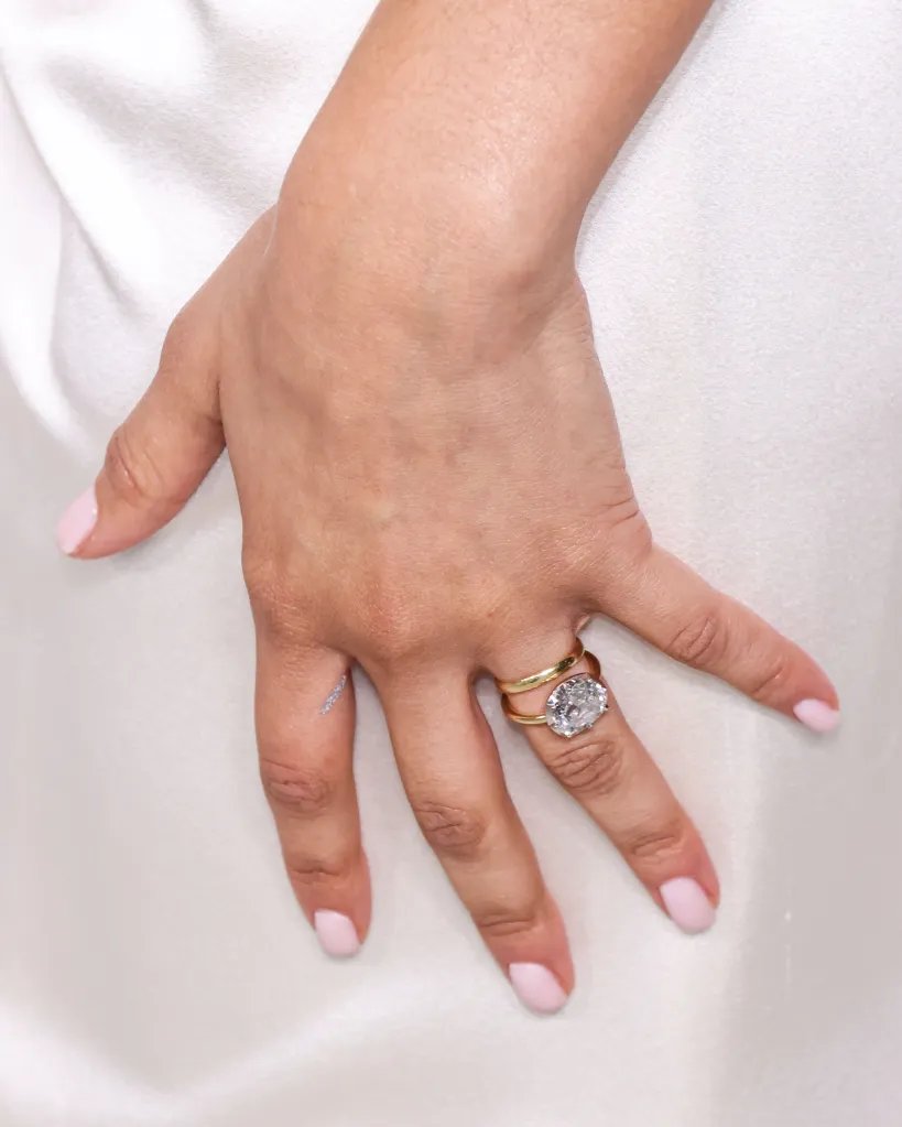 Close-up of Zendaya's hand with light pink manicured nails, wearing a large diamond ring with a thin gold band over a thicker gold band.