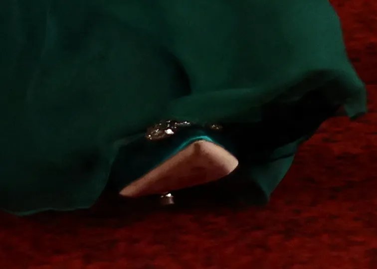 Catherine, Princess of Wales's beige high-heeled shoe beneath her dark green gown, with a diamond buckle on the shoe, on a red carpet.