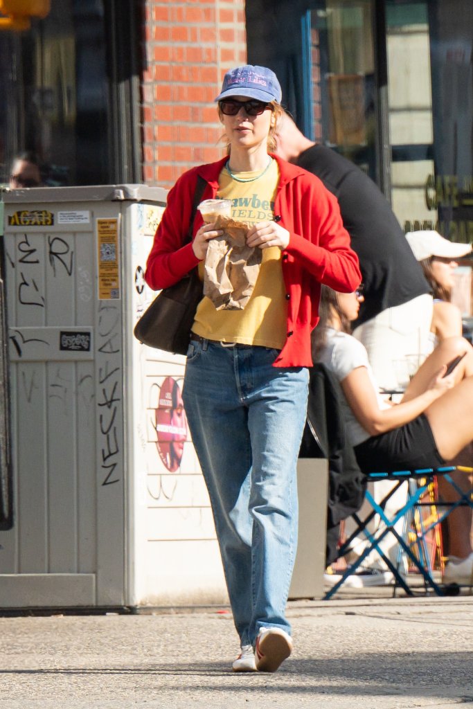 Jennifer Lawrence in a blue cap, sunglasses, red cardigan, yellow t-shirt, blue jeans, and Adidas sneakers, holding coffee and a brown bag in NYC.