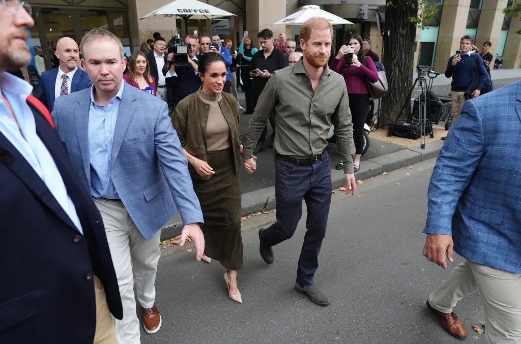 Meghan, Duchess of Sussex, and Prince Harry, Duke of Sussex, walk hand-in-hand while being escorted by security.