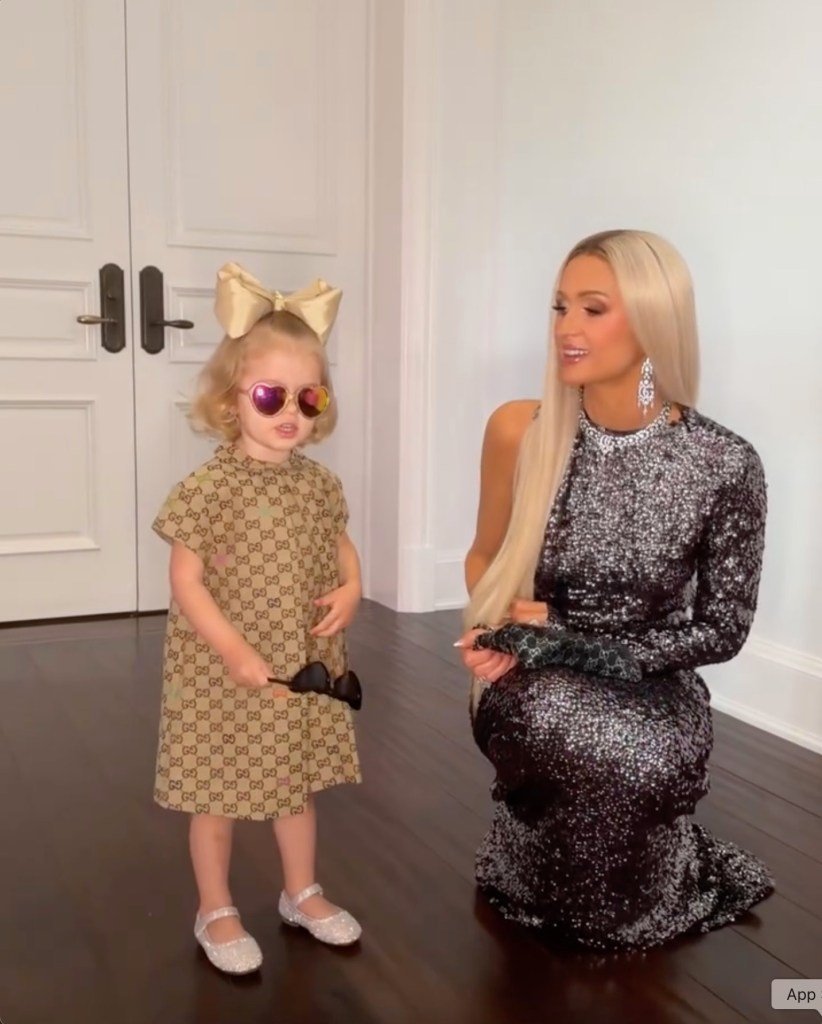 Paris Hilton kneeling in a sequined dress next to a child wearing a Gucci dress, heart-shaped sunglasses, and a large bow.