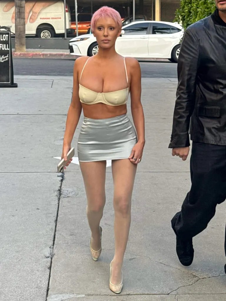 Bianca Censori walking outdoors, wearing a skimpy white bra, shiny silver mini-skirt, sheer stockings, and white heels, with a man in a black jacket partially visible to her right.