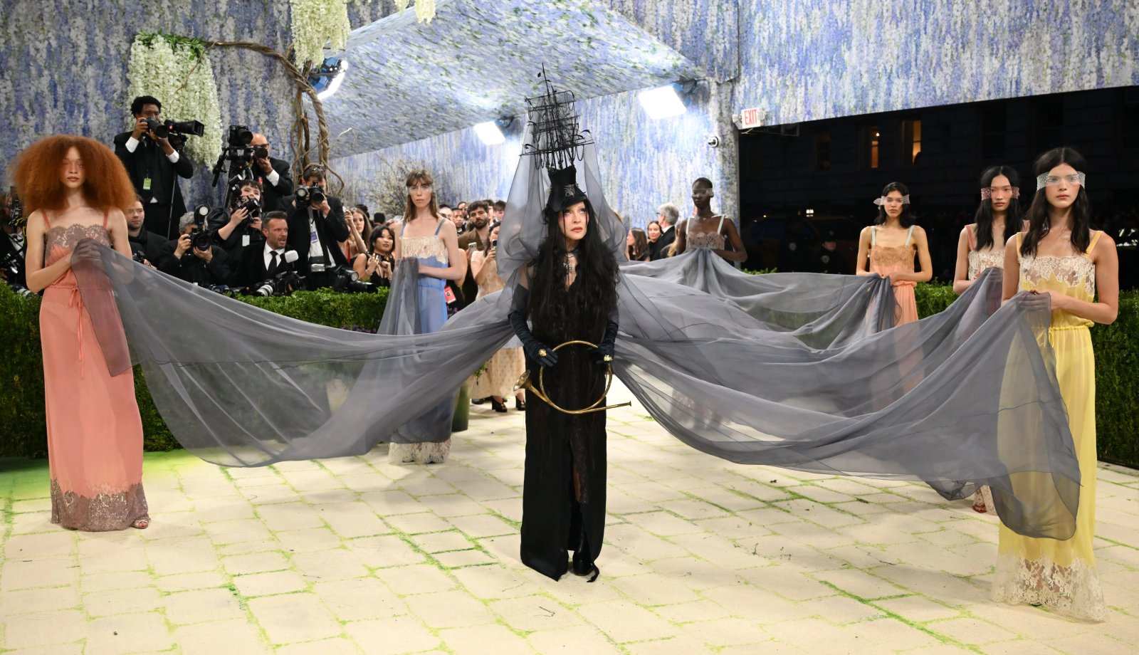 Madonna in a black wig, carrying a horn and wearing a hat resembling a ship, accompanied by models carrying her train at the Met Gala.