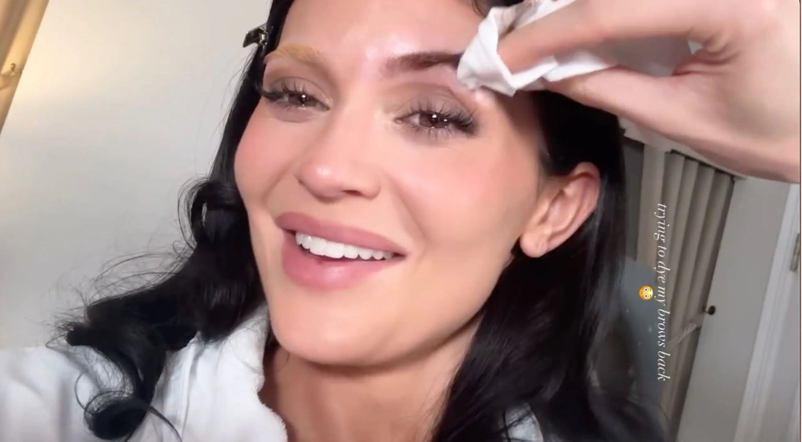 Kylie Jenner in a white robe, smiling while a hand uses a tissue to wipe her eyebrow.