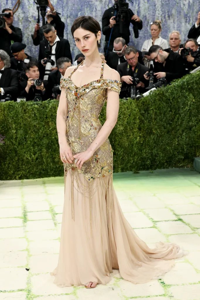 Gracie Abrams in a gold and cream gown with colorful jewels and hanging chains.