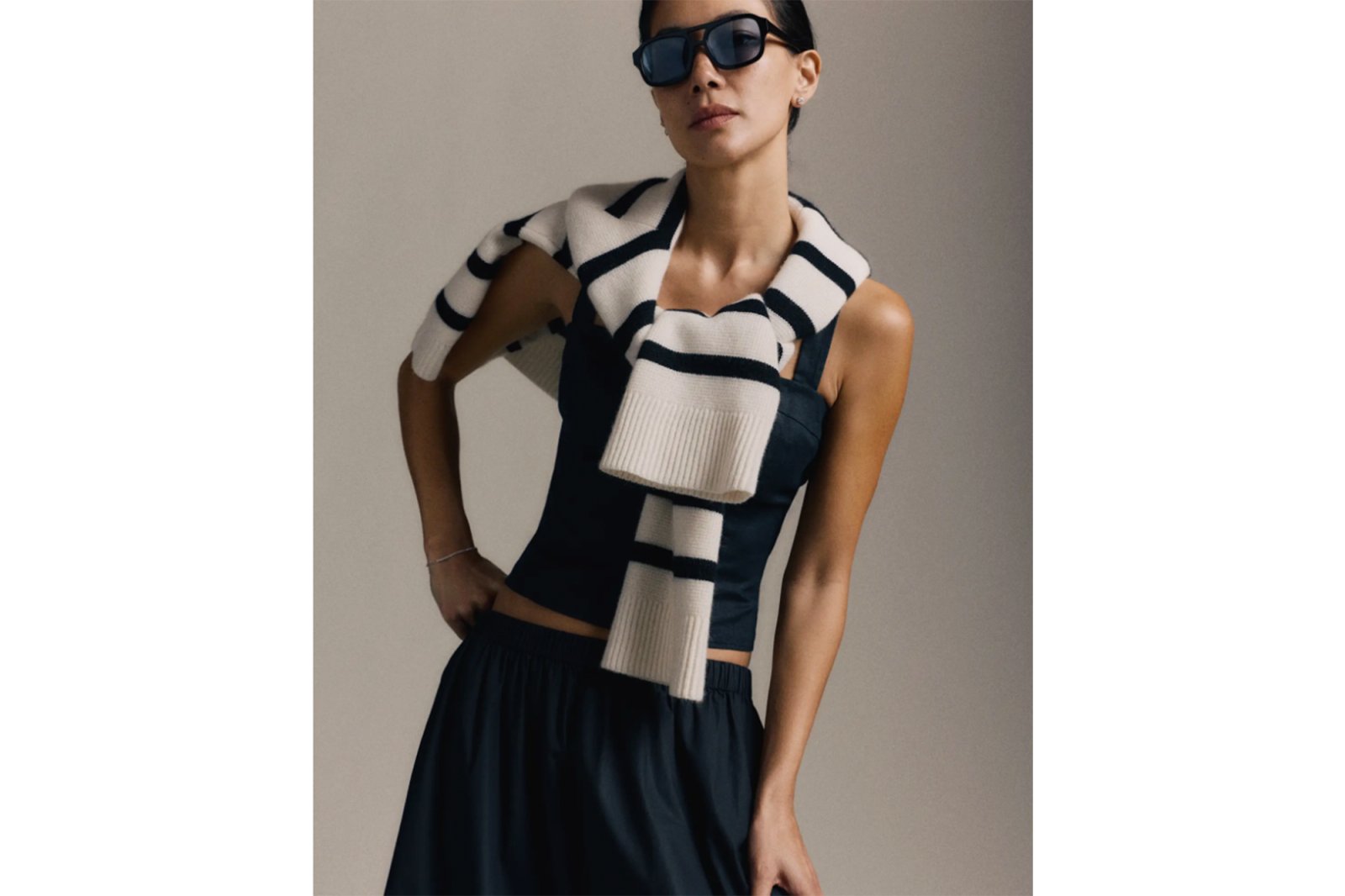 A model in a black top, dark blue skirt, and sunglasses, with a cream and black striped sweater draped over her shoulders.