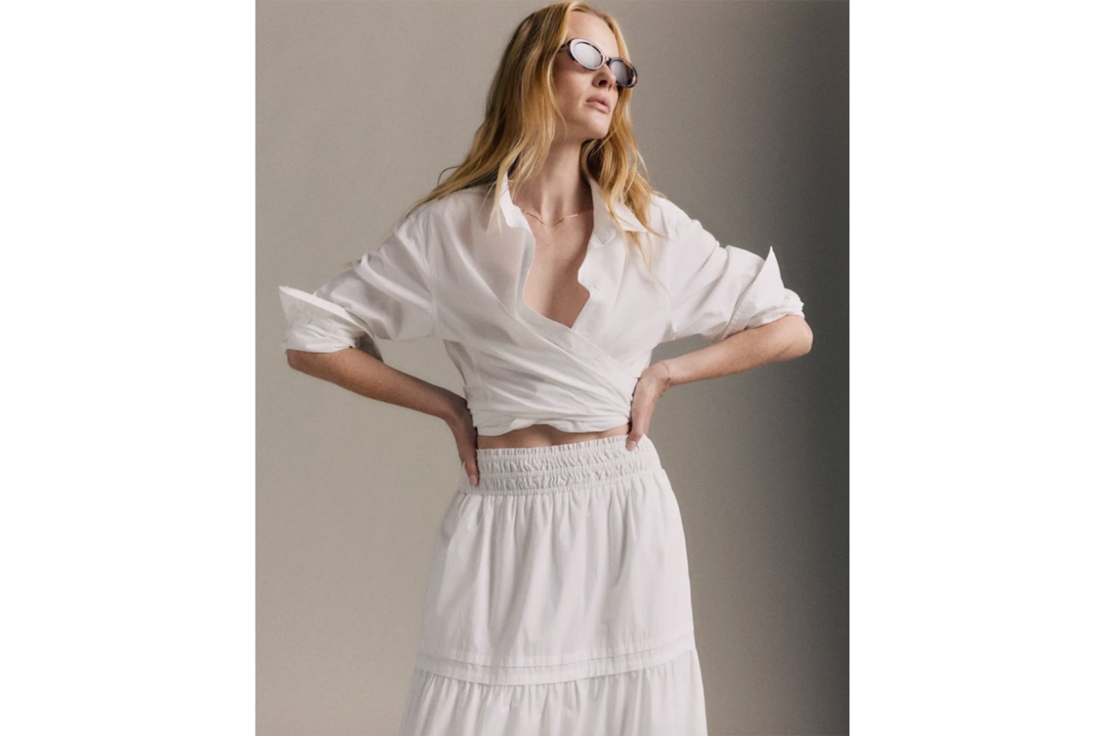 A woman wearing an unbuttoned white shirt tied around her waist and a white tiered skirt, looking up while wearing sunglasses.