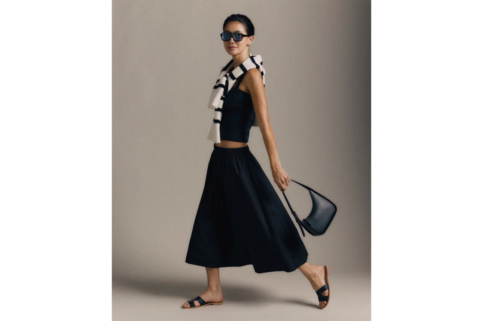 A woman wearing a black crop top, flowing black midi skirt, and black sandals, carrying a purse.