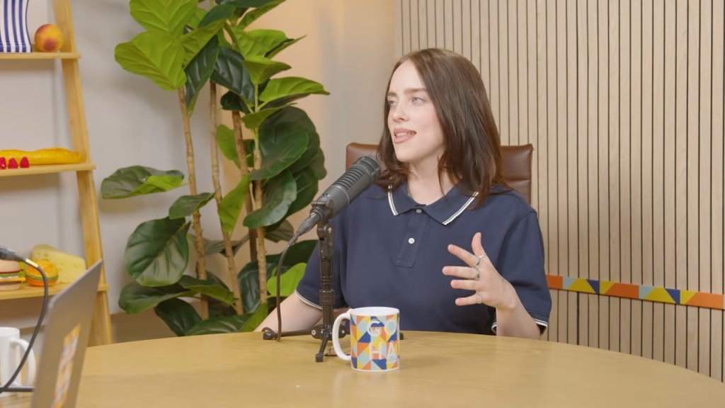 Billie Eilish sitting at a table with a microphone and a mug, gesturing with her left hand.