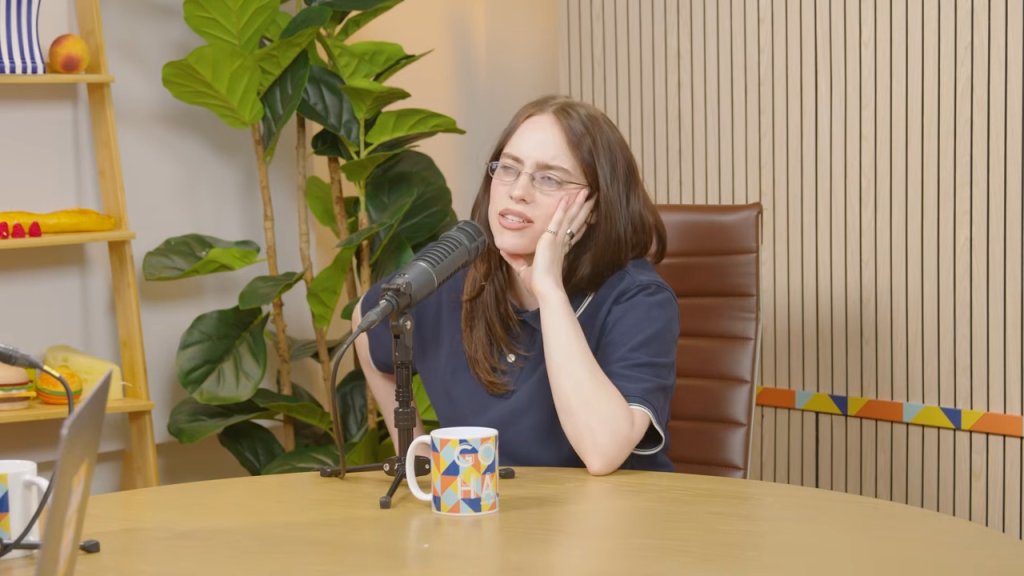 Billie Eilish sitting at a table with a microphone and mug during an interview.