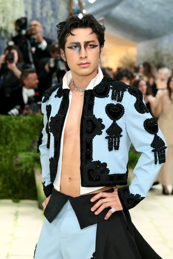 Hudson Williams at the 2026 Met Gala in a light blue jacket with black embroidered details, black pants, and dramatic eye makeup.