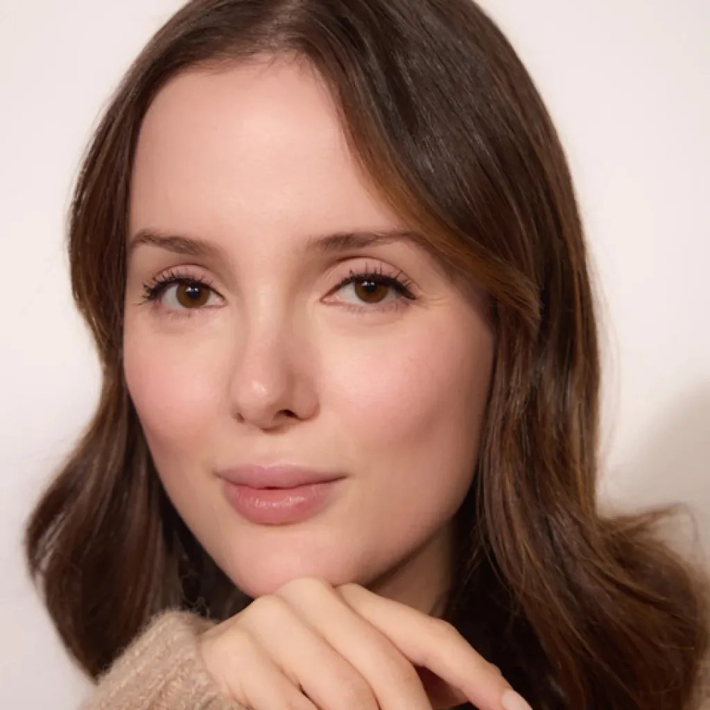 A young woman with brown hair and eyes, chin resting on her hand, wearing a beige sweater.