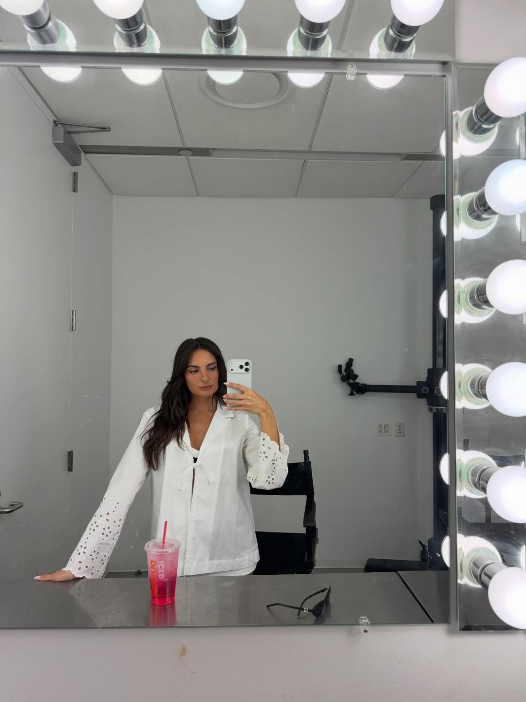 Paige DeSorbo in a white eyelet shirt taking a selfie in a brightly lit dressing room mirror.