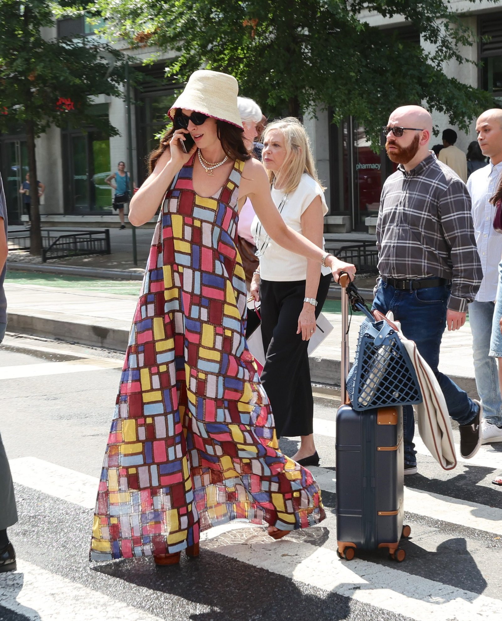 Anne Hathaway in a colorful patterned dress and sunhat, walking with a suitcase while on set for a movie.