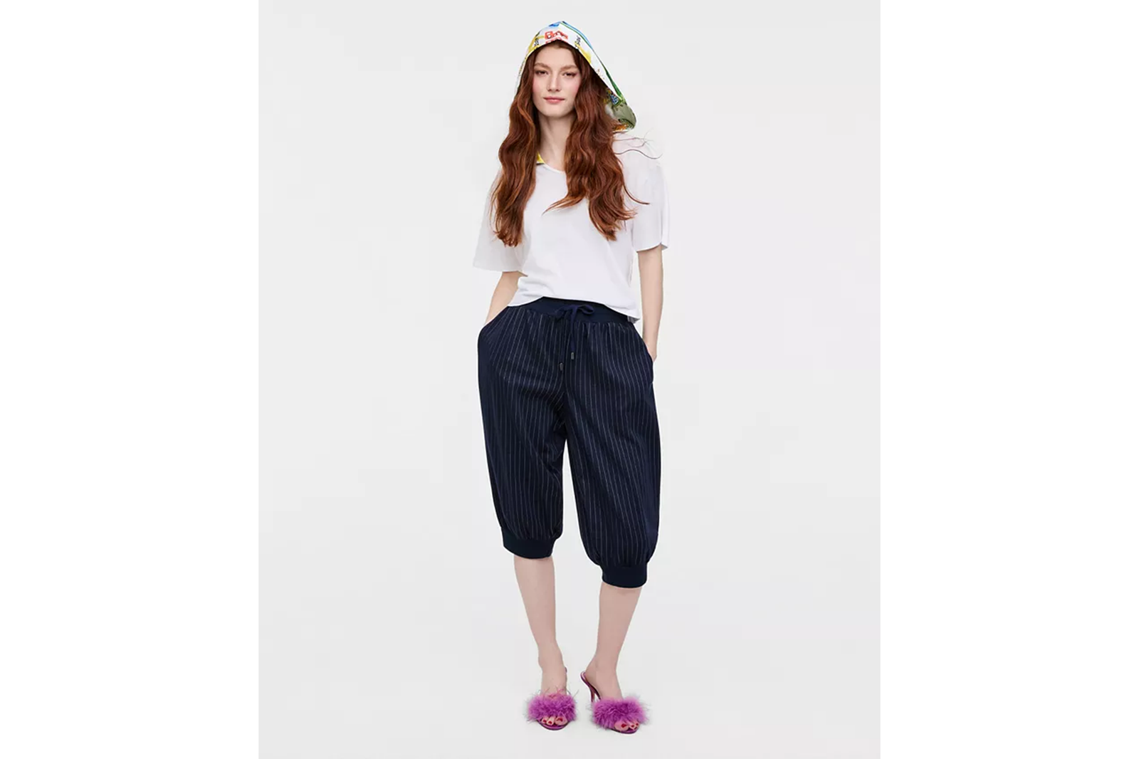 A woman wearing a white t-shirt, dark pinstripe capri pants, purple fluffy heels, and a colorful headscarf.
